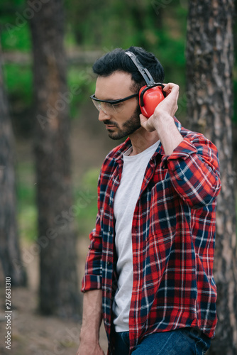 pensive lumberjack in protective glasses standing in forest and touching noise-canceling headphones