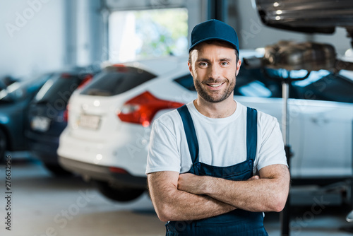 happy car mechanic standing with crossed arms in car service