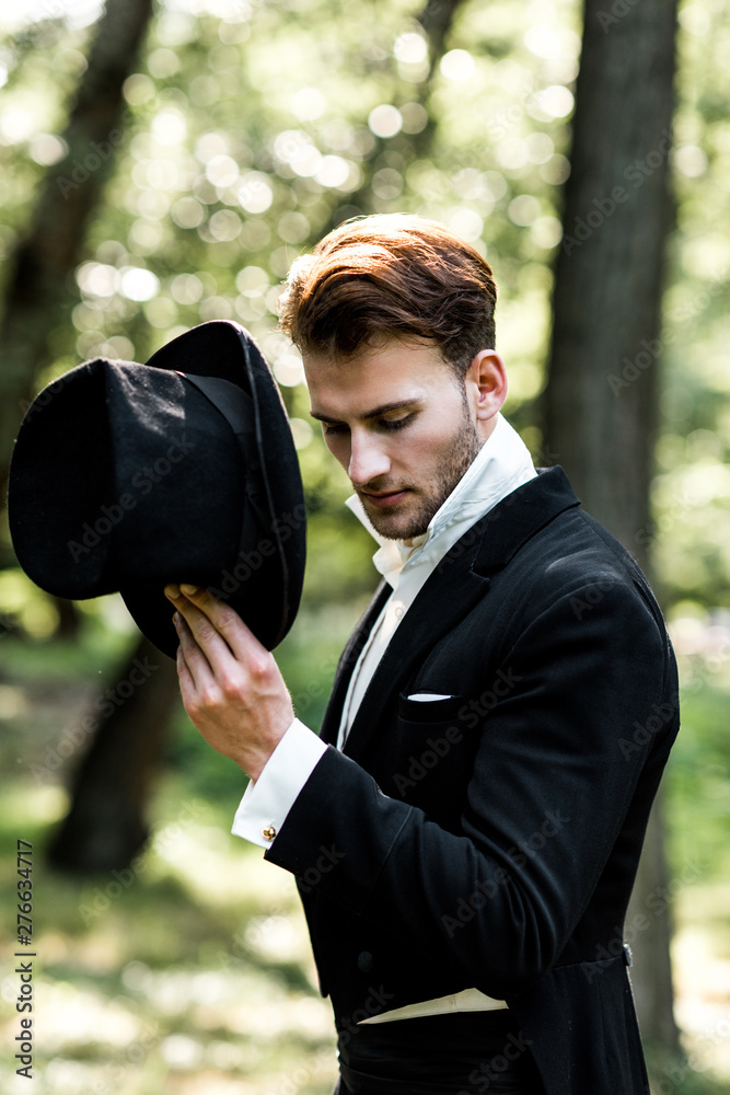 handsome aristocratic man holding hat while standing in suit - obrazy, fototapety, plakaty 