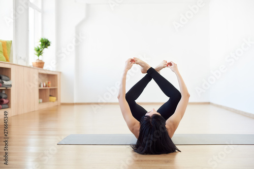 Variation of Happy baby asana. Female brunette, invisible face, lying down on back with hands and legs up doing Ananda Balasana pose, aligning back and stretching spine in light spacious yoga studio