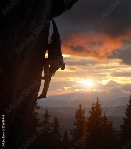 Side view of climber male silhouette in action on steep cliff while shining brightly sun. Rock climbing in nightfall. Beautiful mountains landscape and amazing sunset sky on background. Copy space