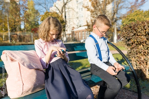 Smart serious children boy and girl are looking into smartphones. On a bench with school backpacks