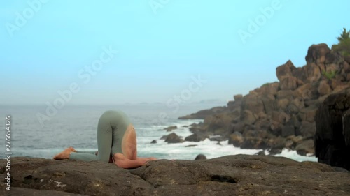 flexible lady stands in karnapidasana on rocky ocean coast against clear sky in calm early morning extreme slow motion. Concept yoga meditation and spiritual practices photo