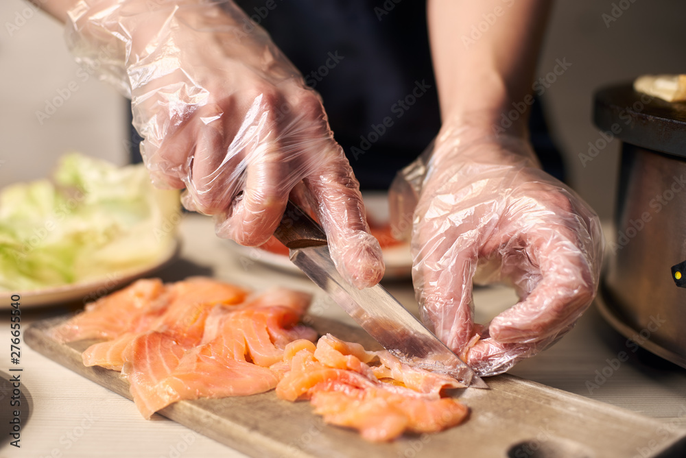 Focus on process of slicing with knife pieces of fish on cutting board for cooking food. Cook's hands in disposable gloves preparing components for dish. Equipment, ingredients on blurred background.