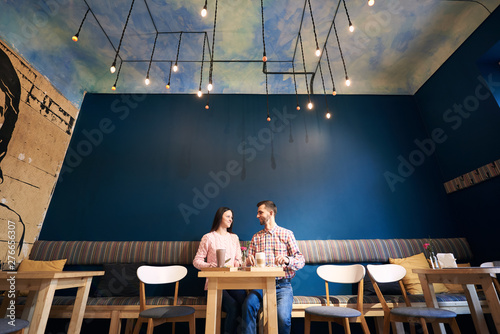 Young couple sitting in atmospheric cafe with interesting ceiling by dating indoors. Full length shot of a male and female student having dinner and talking in cafe isolated on blue background