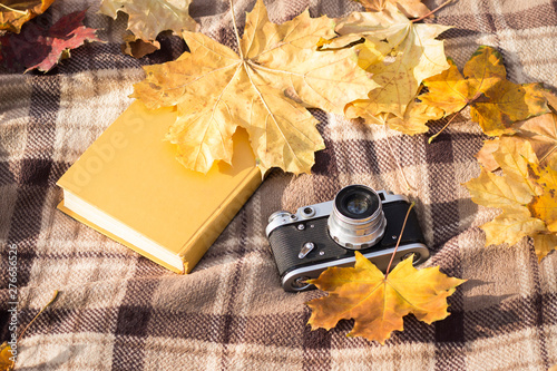 Retro camera and book lay on a brown checkered plaid with yellow autumn leaves. Concept of the fall and outdoor recreation. Flat lay, top view