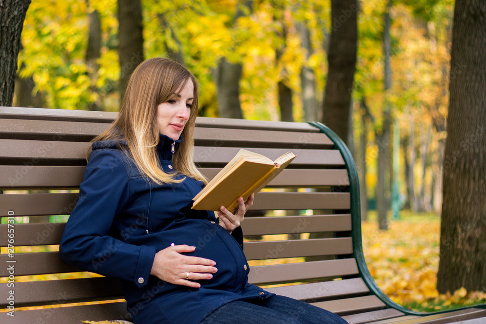 Obraz premium Pregnant young girl sits on a bench and reads a book in an autumn park. Concept relaxing in a park and outdoor