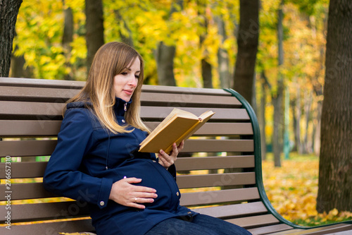 Pregnant young girl sits on a bench and reads a book in an autumn park. Concept relaxing in a park and outdoor