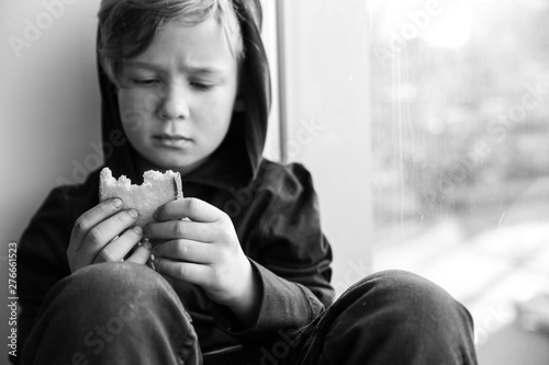 Homeless little boy with bread sitting on window sill indoors