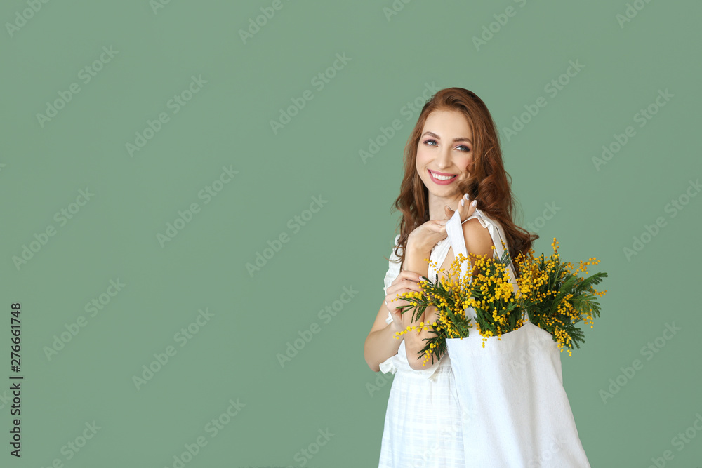 Beautiful young woman holding bag with mimosa flowers on color background