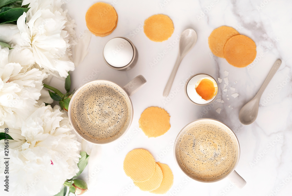 Soft-boiled egg with liquide orange yolk in ceramic egg cups, two cups of coffee with ceramic spoons on background of beautiful white peony. Breakfast concept. Top view. Soft-boiled egg with liquide orange yolk in ceramic egg cups, two cups of coffee with ceramic spoons on background of beautiful white peony. Breakfast concept. Top view.