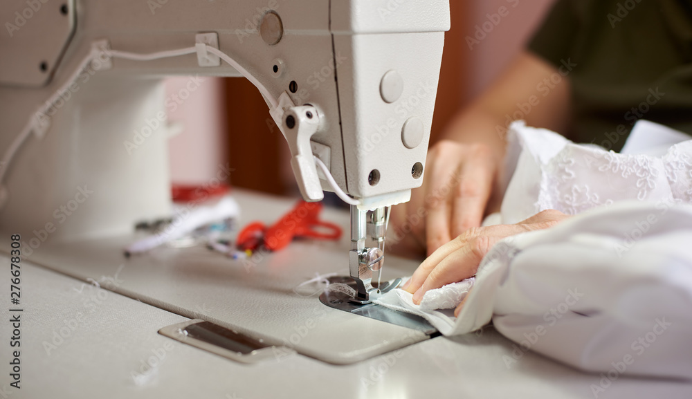 Female hands stitching white fabric on professional manufacturing machine at workplace. Seamstress hands holding lace textile for dress production. Blurred background. Close up view of sewing process.