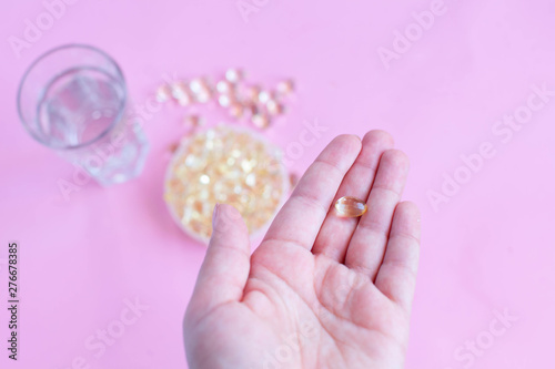 one capsule of omega three on the female palm on a pink background with a glass of water and fish oil top view