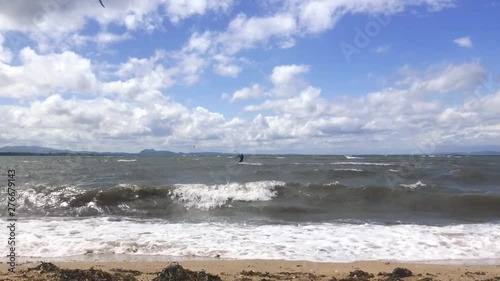 A fixed shot of waves breaking on a sandy beach with kite surfers moving through the shot in the background, on a windy summer day | Portobello beach, Edinburgh | Shot in HD at cinematic 24 fps