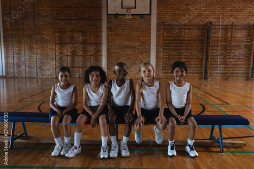 Schoolkids looking at camera while sitting on bench at basketball court