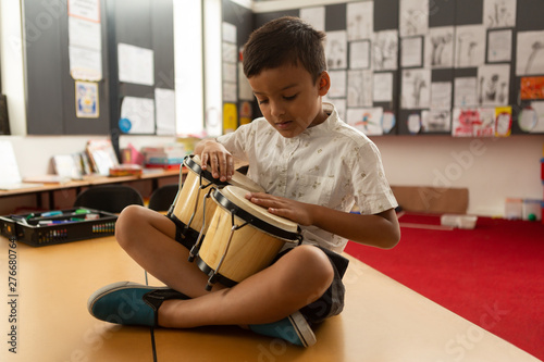 Fotografie Schoolboy sitting on desk and playing bongo in a classroom