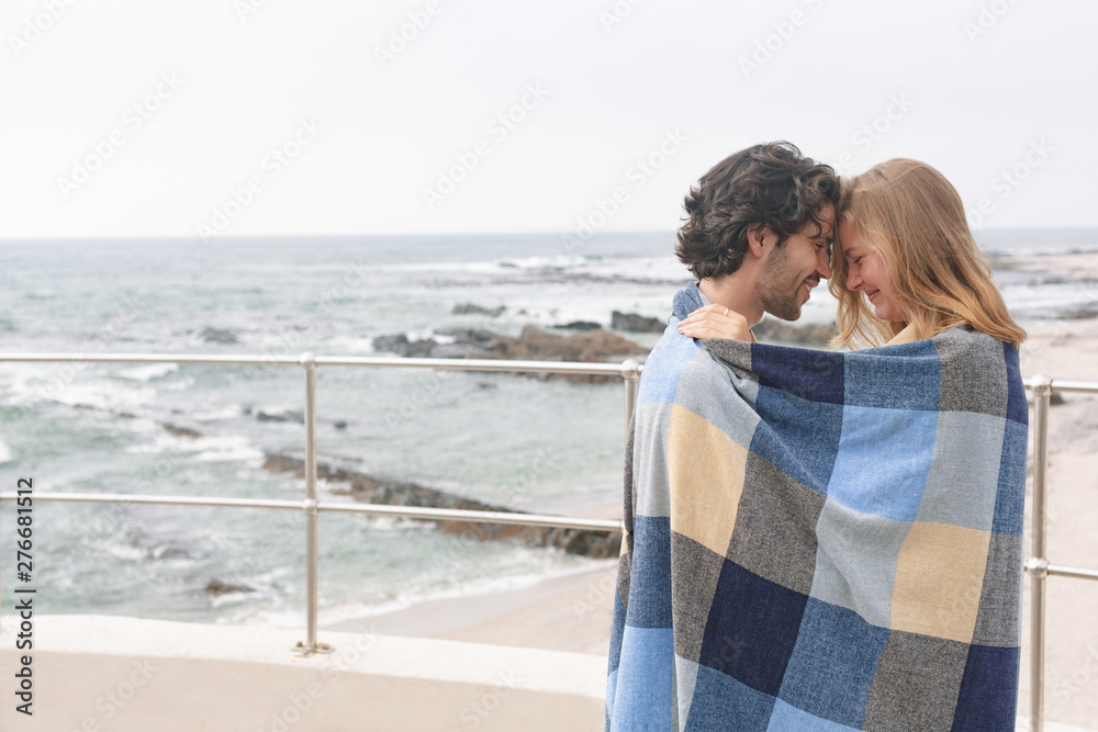 Fototapeta premium Caucasian couple wrapped in blanket standing at promenade near beach 