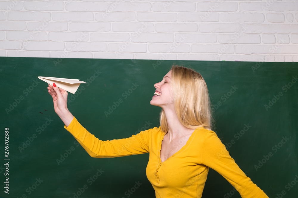 Schoolgirl launches paper plane near blackboard. Origami airplane. Happy girl throwing paper airplane. Girl near school board with paper airplane. Female student playing paper airplane in classroom. Schoolgirl launches paper plane near blackboard. Origami airplane. Happy girl throwing paper airplane. Girl near school board with paper airplane. Female student playing paper airplane in classroom.