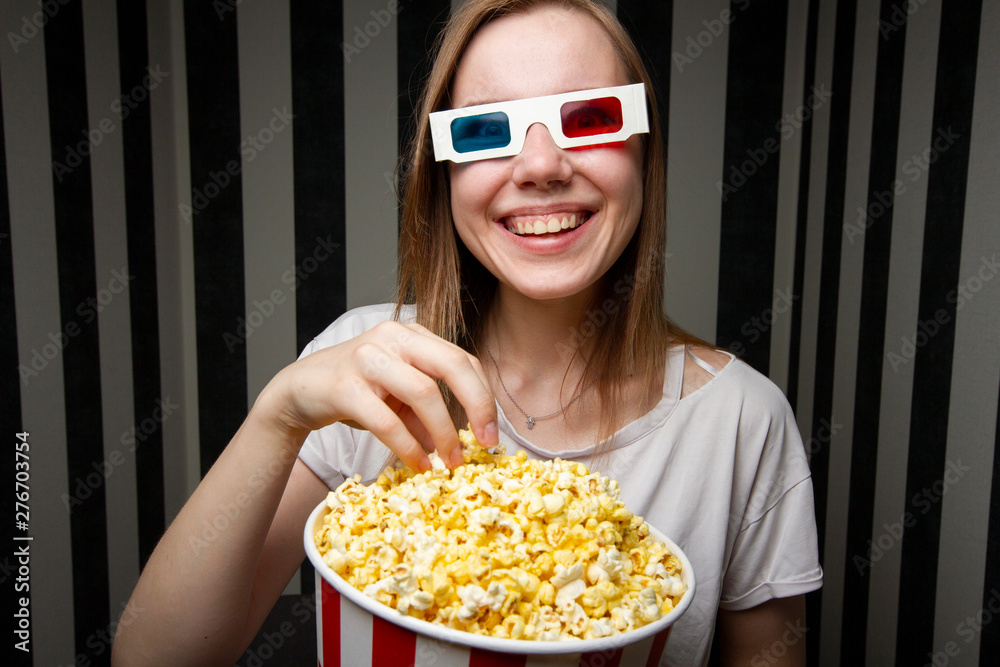 young girl watching a movie and eating popcorn wearing 3d glasses against a striped wall at night, she is emotional and surprised young girl watching a movie and eating popcorn wearing 3d glasses against a striped wall at night, she is emotional and surprised
