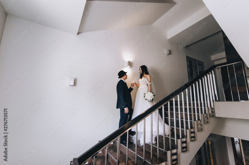 Stylish groom in a blue suit and black hat and a beautiful bride in a white dress are standing on an old staircase. Wedding portrait of newlyweds in love. Concept and photography.