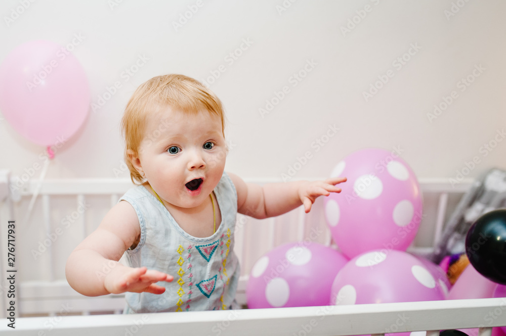Portrait little cute baby girl princess infant 1-2 year standing and playing on bed at home decorated colored balloons for birthday party. Happy baby for a holiday. Celebration first year concept.