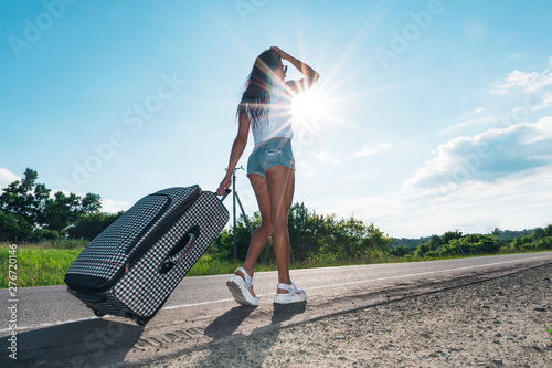 Girl in the bright sun on the road with Luggage on the background of the car. woman go on road with a suitcase. The concept of adventure and travel alone. Road trips. hitchhiking. Summer weekend.