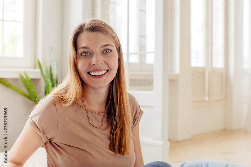 Happy casual adult woman with wide smile sitting in light room looking at camera Happy casual adult woman with wide smile sitting in light room looking at camera