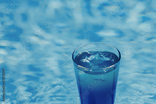 Blue coctail with ice in the glass on the swimming pool background.