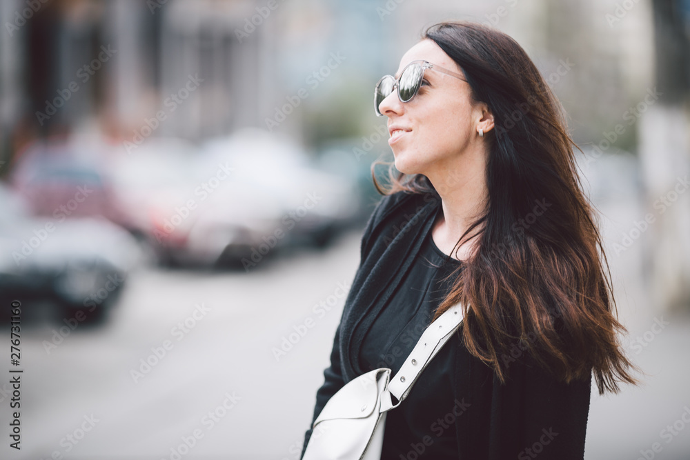 Portrait of a young beautiful caucasian woman on the background of the street. Model girl with long hair and sunglasses with reflection posing in black clothes in an urban environment