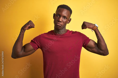 Young african american man wearing red t-shirt standing over isolated yellow background showing arms muscles smiling proud. Fitness concept.