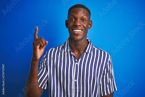 African american man wearing striped casual shirt standing over isolated blue background pointing finger up with successful idea. Exited and happy. Number one.
