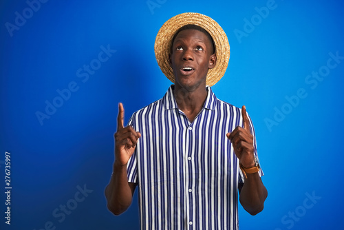 African american man wearing striped shirt and summer hat over isolated blue background amazed and surprised looking up and pointing with fingers and raised arms.