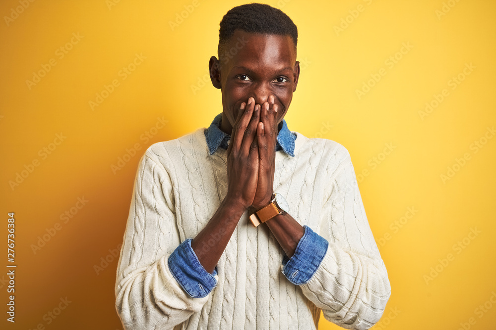African american man wearing denim shirt and white sweater over isolated yellow background laughing and embarrassed giggle covering mouth with hands, gossip and scandal concept African american man wearing denim shirt and white sweater over isolated yellow background laughing and embarrassed giggle covering mouth with hands, gossip and scandal concept