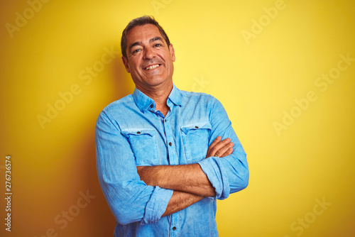 Handsome middle age man wearing denim shirt standing over isolated yellow background happy face smiling with crossed arms looking at the camera. Positive person. © Krakenimages.com
