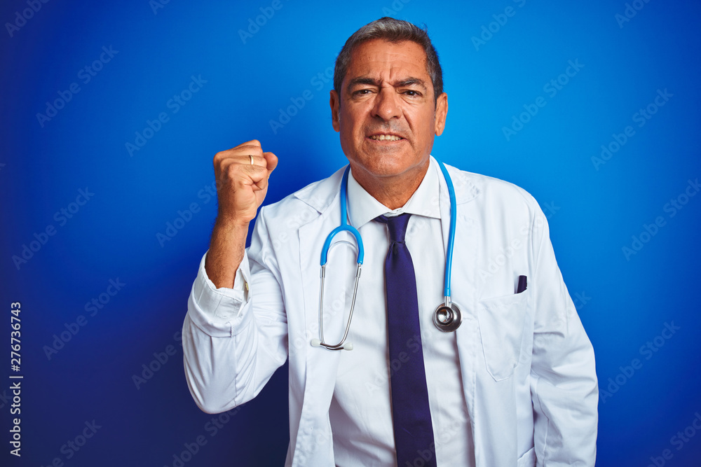 Handsome middle age doctor man wearing stethoscope over isolated blue background angry and mad raising fist frustrated and furious while shouting with anger. Rage and aggressive concept. Handsome middle age doctor man wearing stethoscope over isolated blue background angry and mad raising fist frustrated and furious while shouting with anger. Rage and aggressive concept.