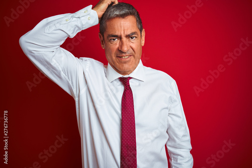 Handsome middle age businessman standing over isolated red background confuse and wonder about question. Uncertain with doubt, thinking with hand on head. Pensive concept.