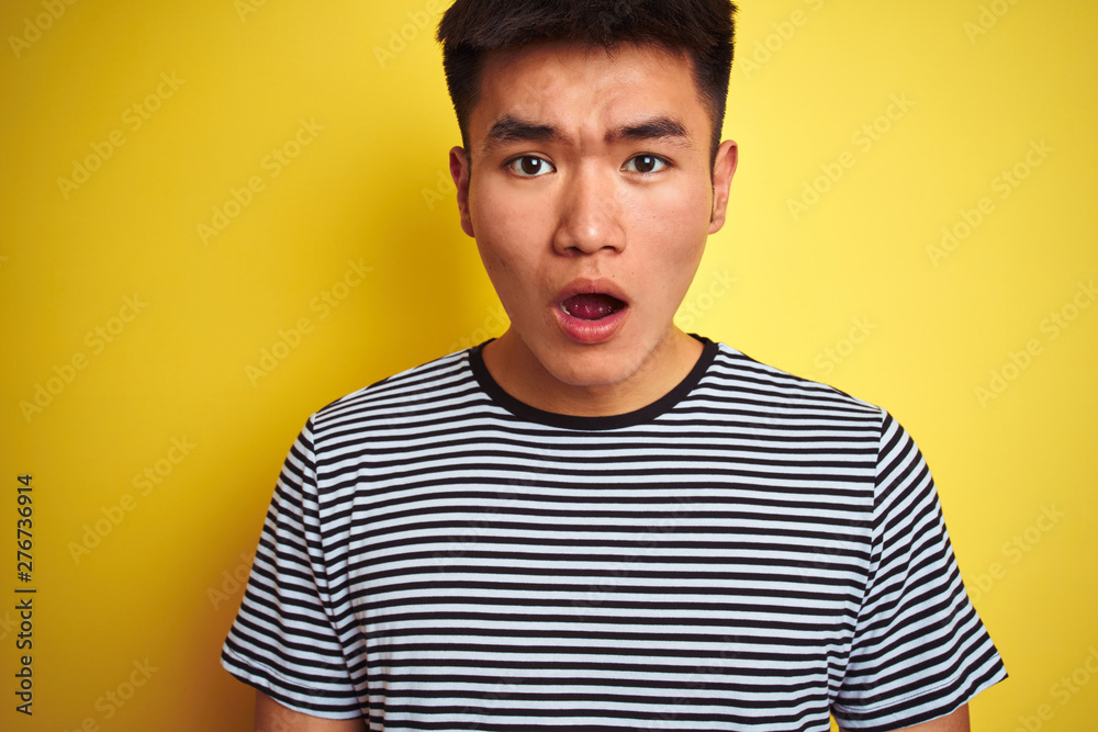Young asian chinese man wearing striped t-shirt standing over isolated yellow background scared in shock with a surprise face, afraid and excited with fear expression Young asian chinese man wearing striped t-shirt standing over isolated yellow background scared in shock with a surprise face, afraid and excited with fear expression