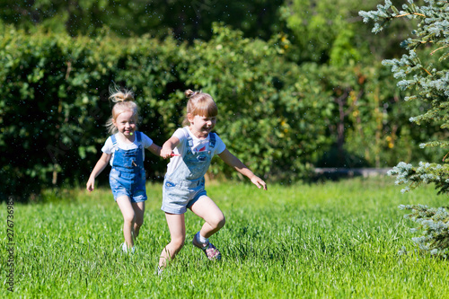 Children run on the green grass holding hands. Girls running around the lawn with grass playing with splashes water to water the plants.