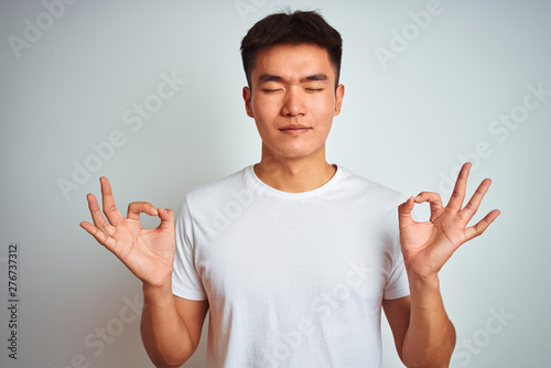 Young asian chinese man wearing t-shirt standing over isolated white background relaxed and smiling with eyes closed doing meditation gesture with fingers. Yoga concept.