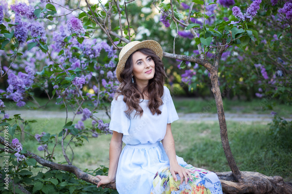 Fototapeta premium Happy Woman Life Style, portrait of a beautiful girl in a straw hat sitting near a blossoming lilac in the rays of the setting sun, picnic. Summer picnic and weekend break concept. Vintage fashion