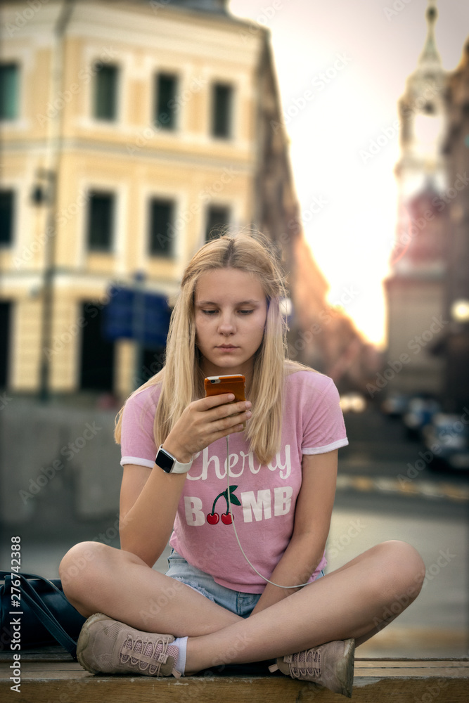 Teen girl sitting in a pink t-shirt on a bench in sunset city, with a phone in her hands.