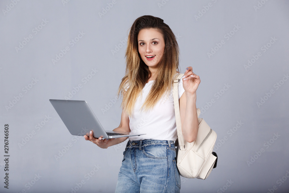 Young sportswoman looks at camera while shopping at the online store. Happy young woman in casual clothes with backpack looking at laptop on gray concrete wall background