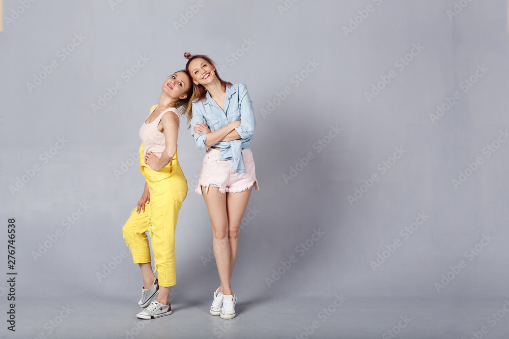 Two beautiful young sisters in stylish clothes are fooled posing in the studio. Beautiful stylish hipsters sisters happily behave against the gray wall background Two beautiful young sisters in stylish clothes are fooled posing in the studio. Beautiful stylish hipsters sisters happily behave against the gray wall background