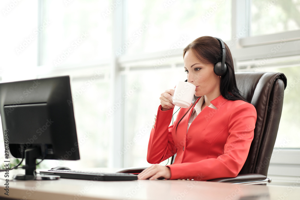 Young attractive woman in red jacket sitting on desk at head of executive in red jacket. Holds video conference in headphones with microphone. Drinking coffee from white cup. Young attractive woman in red jacket sitting on desk at head of executive in red jacket. Holds video conference in headphones with microphone. Drinking coffee from white cup.