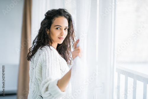 Young woman opening window curtains at home.