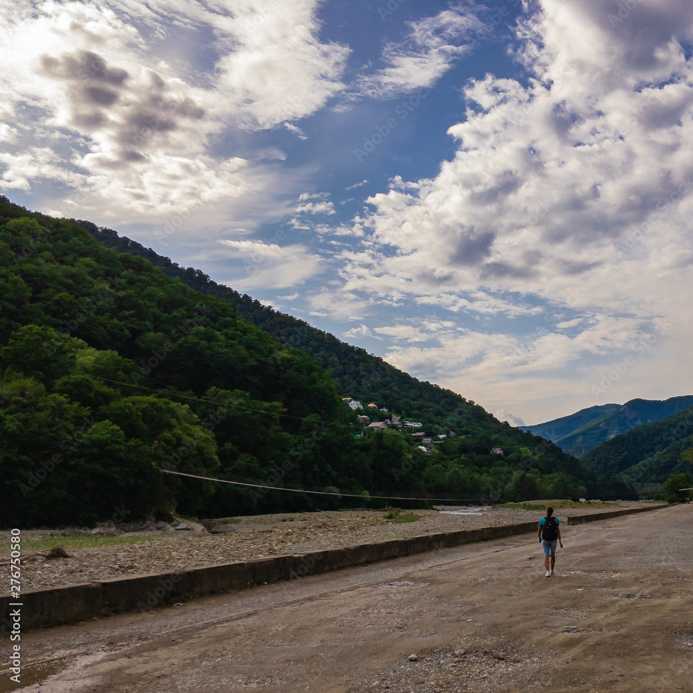 A man with a backpack is walking along the road towards the mountains on a summer day.