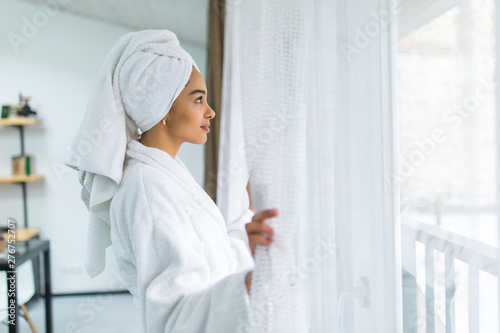 Beautiful young woman in white robe and towel on head standing near window and open curtains in the morning after shower