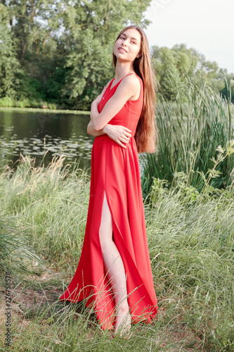 Brown-haired barefoot girl with long hair in red dress on river coast.