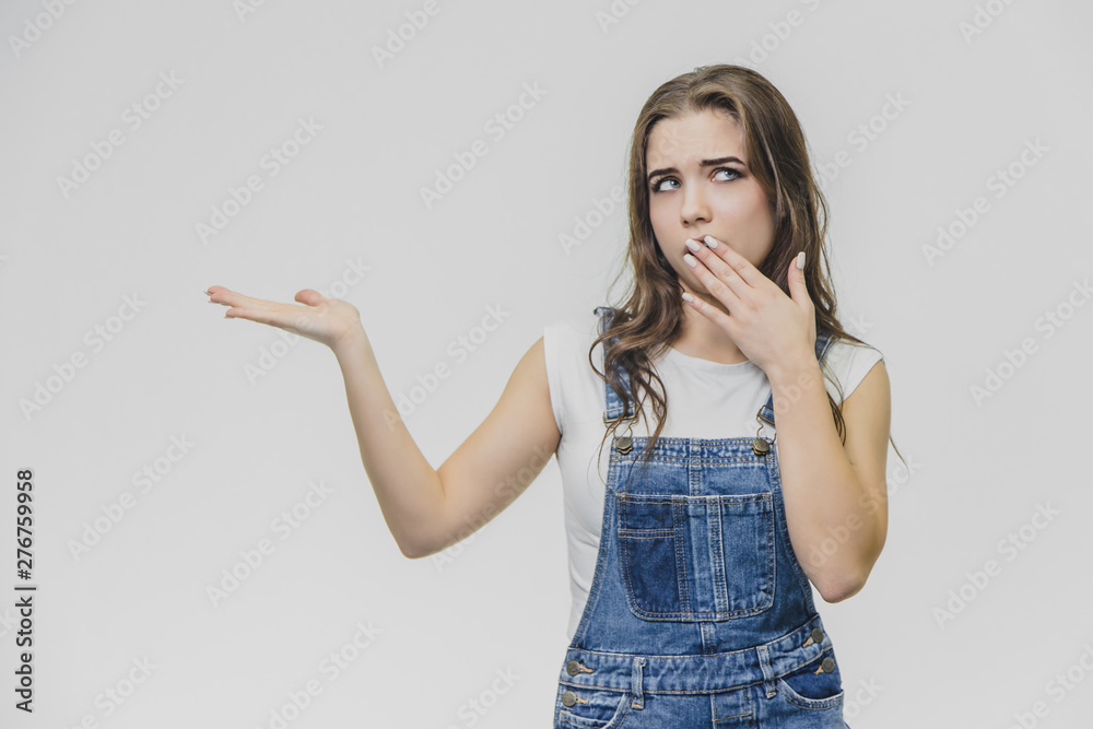 Young student girl standing on a white background. Dressed in white t-shirt and denim overalls. During this time, putting one hand to one side, the other hand shields his mouth and looks at the camera Young student girl standing on a white background. Dressed in white t-shirt and denim overalls. During this time, putting one hand to one side, the other hand shields his mouth and looks at the camera