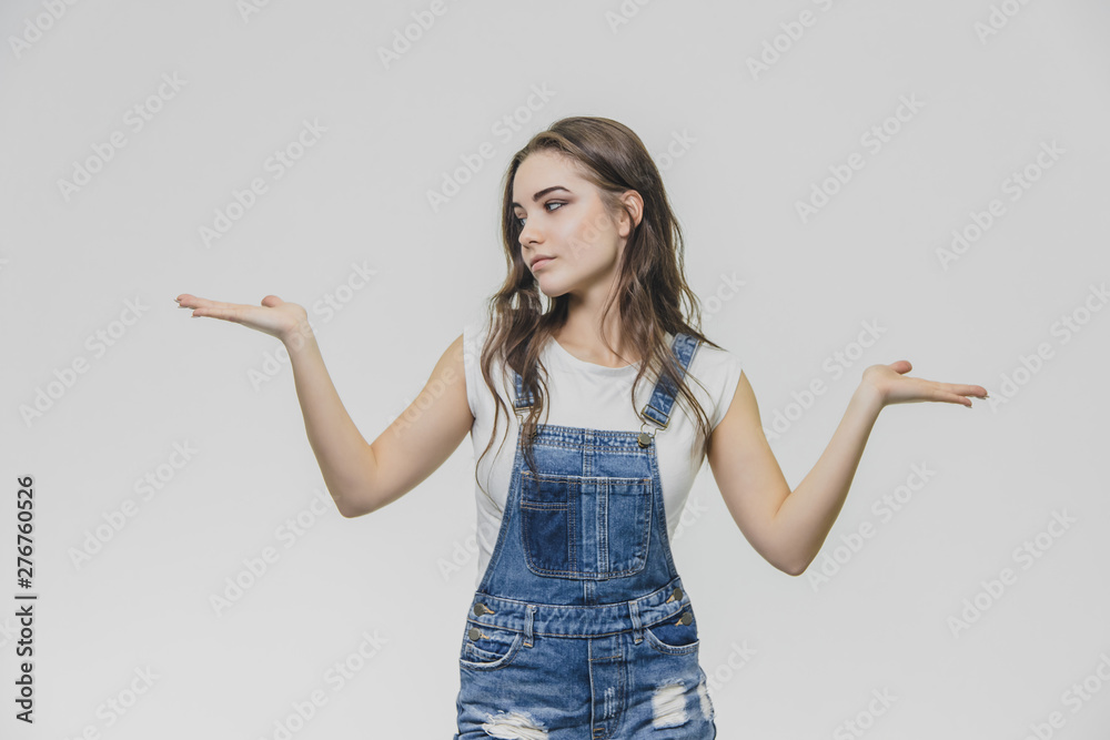 Happy brunette teenager standing on gray background. Dressed in a denim overalls and a white T-shirt. Looking side by side the girl put her hands in different directions, surprised.